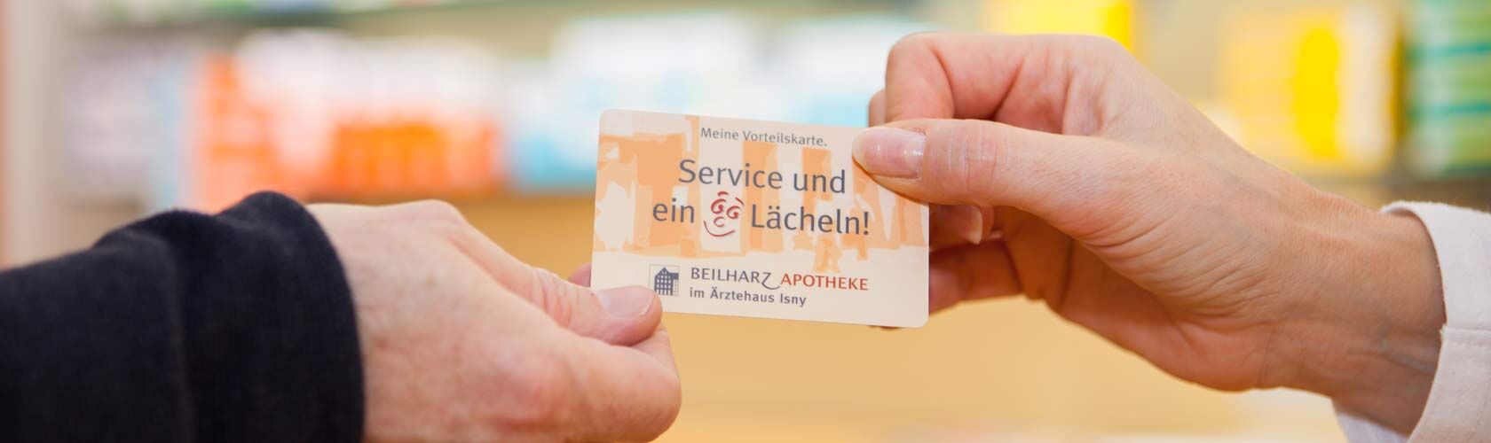 Zwei Personen tauschen eine Servicekarte der Beilharz Apotheke Isny mit dem Slogan "Service und ein Lächeln" aus.